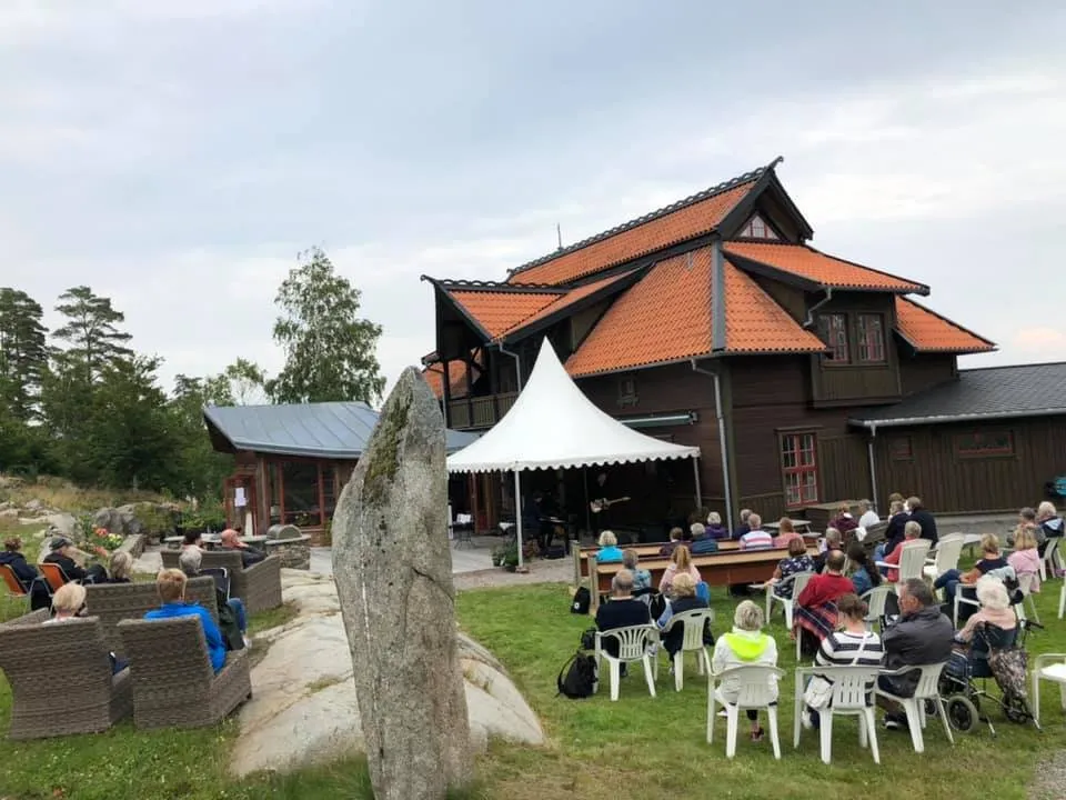 Publik vid borden på gräsmattan framför huset, med ett vitt tält och en rest sten